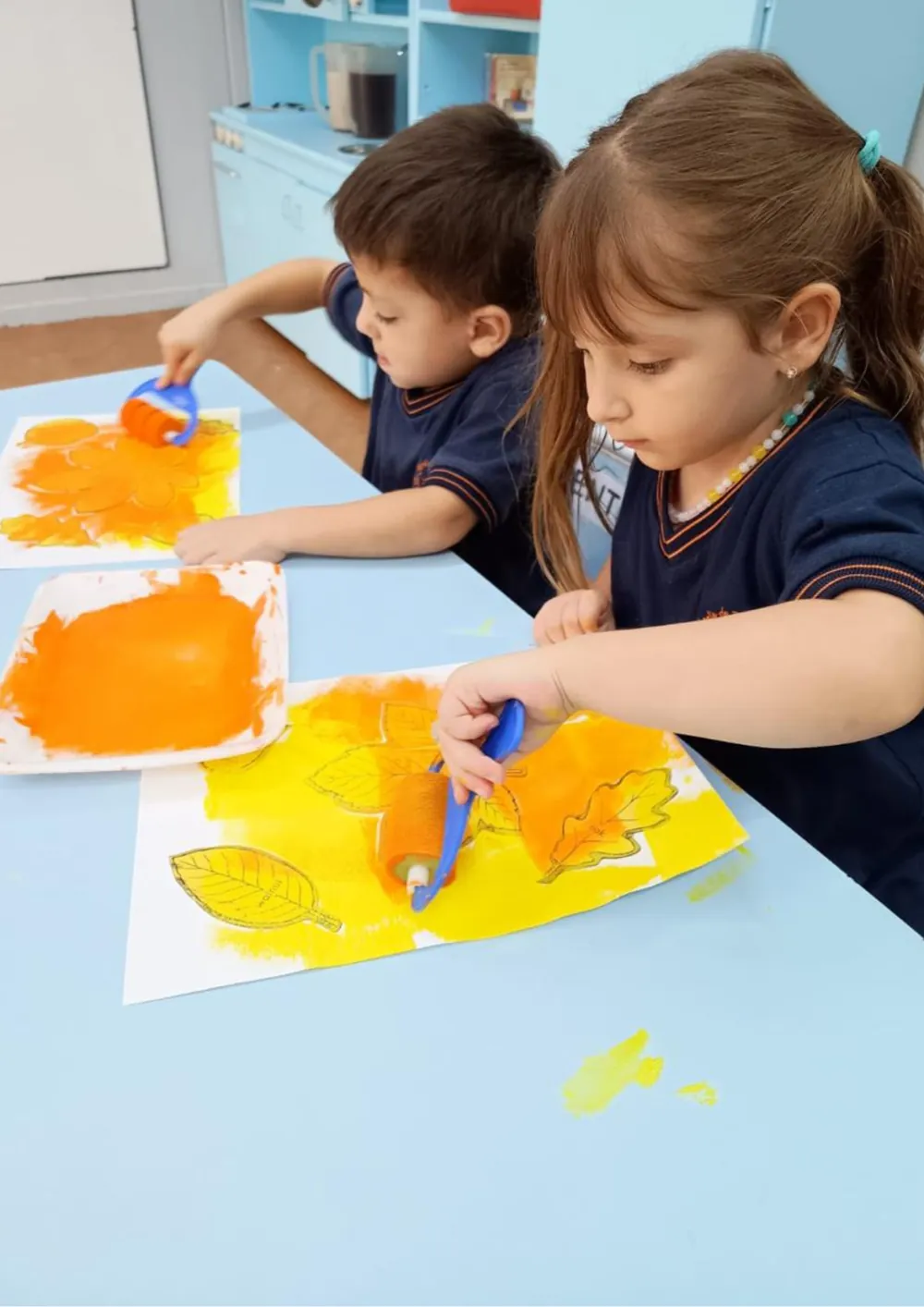 Dos niños del jardín pintando hojas con rodillos y témpera naranja